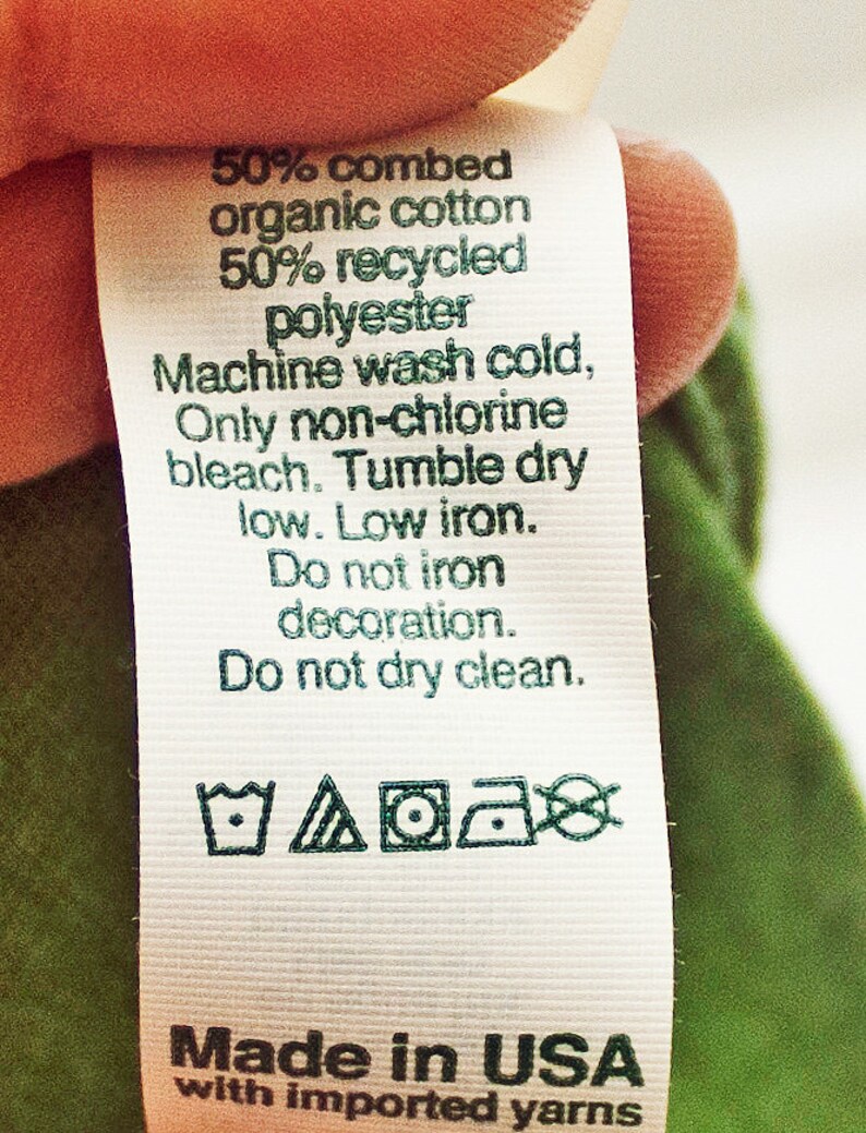 May include: White care label with green text and symbols. The label states "50% combed organic cotton 50% recycled polyester. Machine wash cold, Only non-chlorine bleach. Tumble dry low. Low iron. Do not iron decoration. Do not dry clean." The label also includes four symbols: a tub of water with a wavy line, a triangle with a circle inside, a circle with a dot inside, and a circle with a diagonal line through it. The label concludes with "Made in USA with imported yarns."