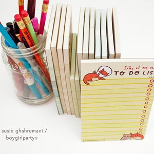 May include: A yellow to-do list notepad with a red cat illustration and the text "like it or not TO DO LIST" on the front. The notepad is sitting on a table with a glass jar filled with colorful pencils and pens.