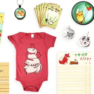 May include: A collection of items featuring cute animal illustrations.  The items include a book titled "What Will Hatch?", a red baby onesie with a stack of cats, a to-do list notepad with a fox, a small notebook with a forest scene, and several small glass ornaments with animal designs.
