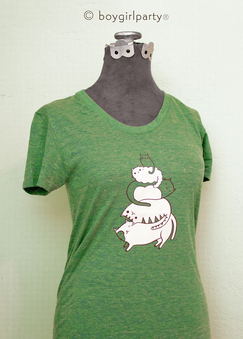 May include: Green t-shirt with a graphic of a stack of cats. The cats are white with brown outlines and are stacked on top of each other. The text "boygirlparty" is printed in brown at the top of the shirt.