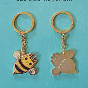 May include: Gold keychain featuring a cartoon bee with a cat face. The bee is yellow and black with a gold crown. The back of the keychain is plain gold.