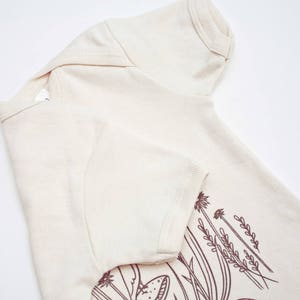 Puede incluir: Una camiseta de manga corta de color crema con un dibujo lineal marr&oacute;n de flores silvestres y setas en la mitad inferior de la camiseta.