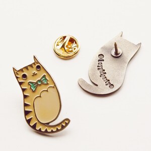 May include: A gold enamel pin of a cartoon cat wearing a green bow tie. The cat is sitting with its tail curled around its body. The back of the pin is silver and says "boygirlparty".
