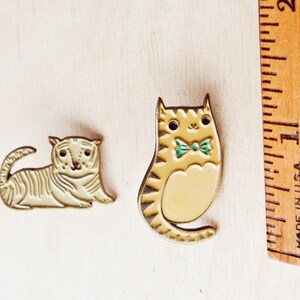 May include: Two enamel pins featuring cartoon cats. One cat is white with black stripes and the other is yellow with black stripes. Both cats have green eyes and a green bow tie.