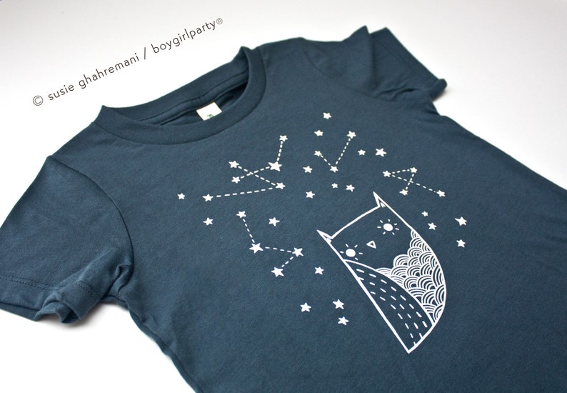 May include: A dark blue t-shirt with a white owl and constellation design. The owl is looking up at the stars. The text "susie ghahremani / boygirlparty&reg;" is in the top left corner.