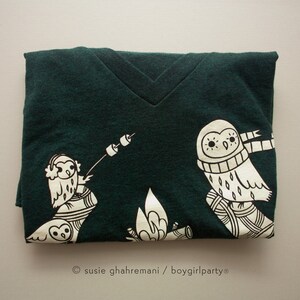 May include: A dark green t-shirt with a white graphic of two owls sitting by a campfire. The owls are wearing scarves and one is holding a stick with a marshmallow on the end. The text "susie ghahremani / boygirlparty&reg;" is printed on the bottom of the shirt.