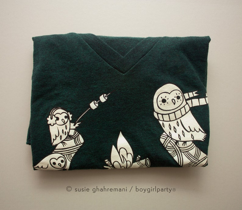 May include: A dark green t-shirt with a white graphic of two owls sitting by a campfire. The owls are wearing scarves and one is holding a stick with a marshmallow on the end. The text "susie ghahremani / boygirlparty&reg;" is printed on the bottom of the shirt.