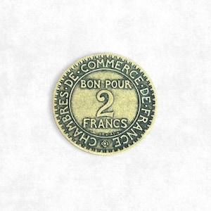 Pode incluir: Uma moeda vintage circular com o texto "BON POUR 2 FRANCS". A moeda tem um esquema de cores verde claro e dourado, com as palavras "CHAMBRES DE COMMERCE DE FRANCE" ao redor da borda. Um objeto de coleção.