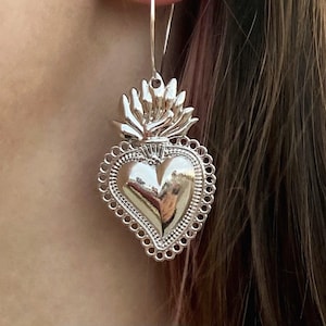 May include: Silver heart-shaped earrings with a detailed design and a flame-like top. The earrings are hanging from a person's ear.