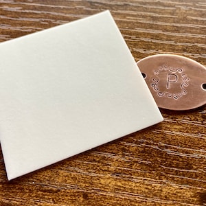 Puede incluir: Una pequeña etiqueta ovalada de cobre con un diseño decorativo y la letra "P" en el centro. La etiqueta tiene dos agujeros en el lateral.