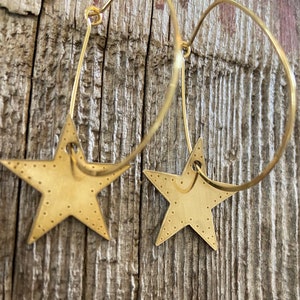 Könnte beinhalten: Ein Paar goldene Creolen mit einem sternförmigen Anhänger, der an jedem Creolen hängt. Die Sterne haben eine strukturierte Oberfläche und sind hellgoldfarben.