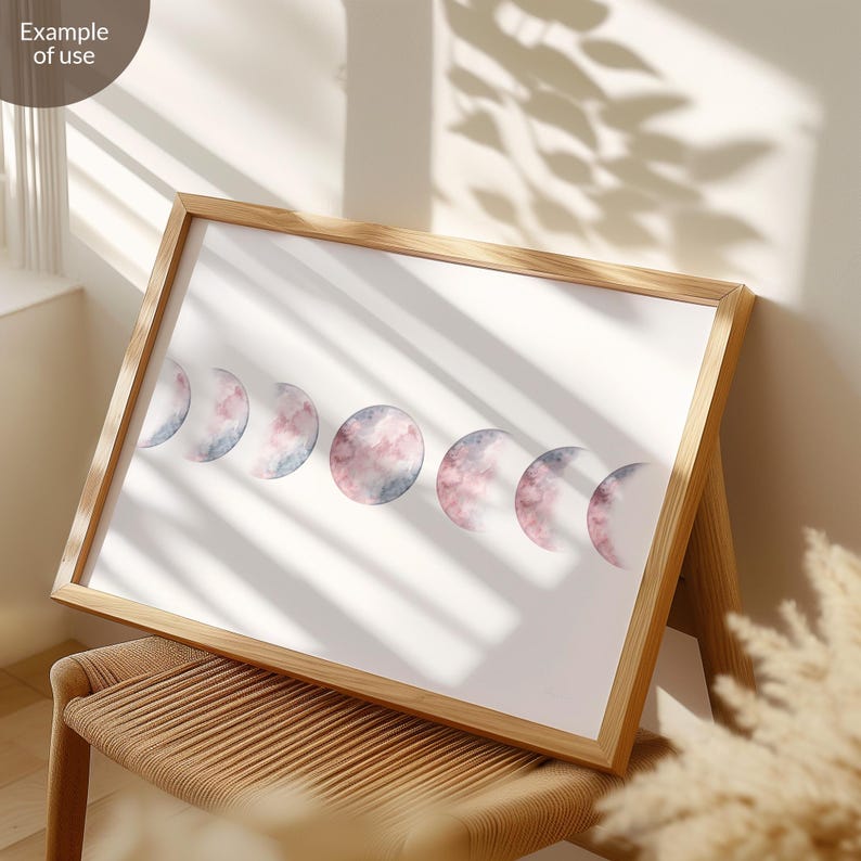May include: A framed print of a watercolor painting depicting the phases of the moon. The moon is painted in shades of pink and blue, with a soft, ethereal look. The print is framed in a natural wood frame.