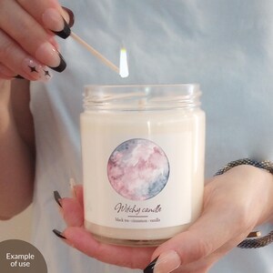 May include: A glass jar candle with a white wax and a label that says "Witchy candle" and "black tea - cinnamon - vanilla". The label also features a watercolor illustration of a pink and blue moon.