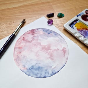 May include: A watercolor painting of a pink and blue moon on white paper. A paintbrush and a palette of paints are visible in the background.