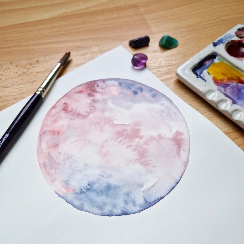 May include: A watercolor painting of a pink and blue moon on white paper. A paintbrush and a palette of paints are visible in the background.