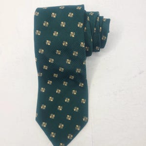 Puede incluir: Una corbata de color verde azulado oscuro con un patrón repetitivo de pequeños motivos cuadrados claros dispuestos en cuadrícula. La tela tiene una sutil textura acanalada diagonal y la corbata está doblada en la parte superior para mostrar su estructura.