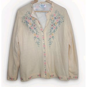 May include: Cream-colored cardigan sweater with floral embroidery in pink, blue, and yellow. The sweater has a button-up front and long sleeves. The floral design is on the front and along the bottom hem.