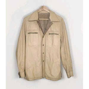 May include: A tan leather jacket with a collared neckline and two zippered chest pockets. The jacket has a button-down front and long sleeves with buttoned cuffs. It is hanging on a wooden hanger against a white background.
