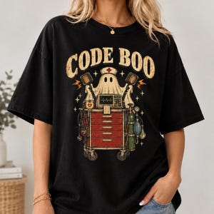 May include: A black t-shirt featuring a graphic print of a ghost wearing a nurse cap, standing behind a red medical crash cart with a heart monitor, oxygen tank, and stethoscope. The text CODE BOO is printed in a distressed beige font above the ghost.
