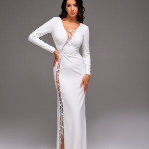Könnte beinhalten: Ein weißes, langärmeliges Maxikleid mit tiefem Ausschnitt und einer Spitzenüberlagerung. Das Kleid hat einen hohen Schlitz auf der linken Seite.