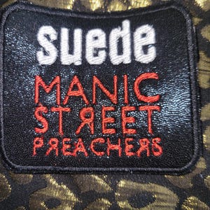 May include: A rectangular black embroidered patch featuring the white text "suede" above the red text "MANIC STREET PREACHERS" with the letters "R" reversed. The patch is set against a black fabric background with a metallic gold leopard print pattern.