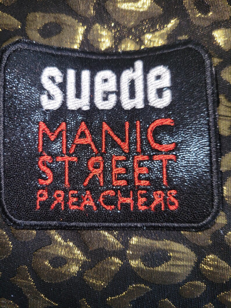May include: A rectangular black embroidered patch featuring the white text "suede" above the red text "MANIC STREET PREACHERS" with the letters "R" reversed. The patch is set against a black fabric background with a metallic gold leopard print pattern.