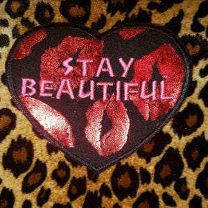 Pode incluir: Um patch em forma de coração com as palavras "STAY BEAUTIFUL" bordadas em rosa. O patch apresenta lábios vermelhos e é colocado sobre um fundo com estampa de leopardo. O patch é preto com um design de lábios vermelhos.
