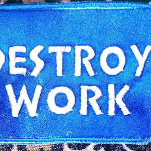 Könnte beinhalten: Ein blaues rechteckiges Patch mit weiß gesticktem Text, der "DESTROY WORK" lautet. Der Aufnäher hat einen gestickten Rand und befindet sich vor einem gemusterten Hintergrund.