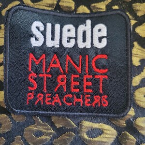 May include: A black rectangular patch with white and red embroidered text reading Suede Manic Street Preachers, featuring a reversed R and E in the word Preachers, stitched onto a fabric with a black and metallic gold leopard print pattern.