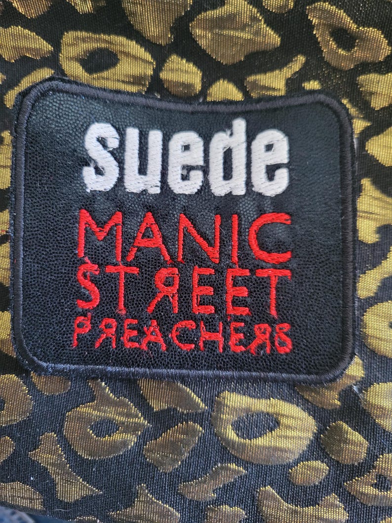May include: A black rectangular patch with white and red embroidered text reading Suede Manic Street Preachers, featuring a reversed R and E in the word Preachers, stitched onto a fabric with a black and metallic gold leopard print pattern.