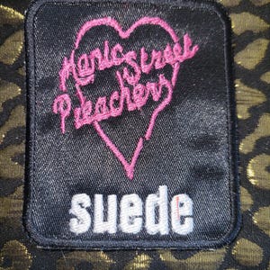 May include: A rectangular black fabric patch featuring an embroidered pink heart outline containing the text Manic Street Preachers in pink thread, with the word suede embroidered in white block letters below the heart, set against a gold leopard print.
