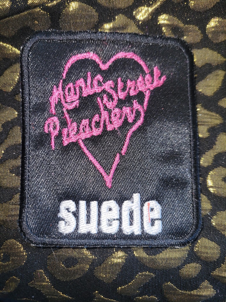 May include: A rectangular black fabric patch featuring an embroidered pink heart outline containing the text Manic Street Preachers in pink thread, with the word suede embroidered in white block letters below the heart, set against a gold leopard print.