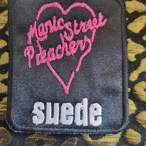 May include: A rectangular black fabric patch featuring an embroidered pink heart outline containing the text Manic Street Preachers in pink script. Below the heart, the word suede is embroidered in white block letters. The patch is on a gold leopard print.
