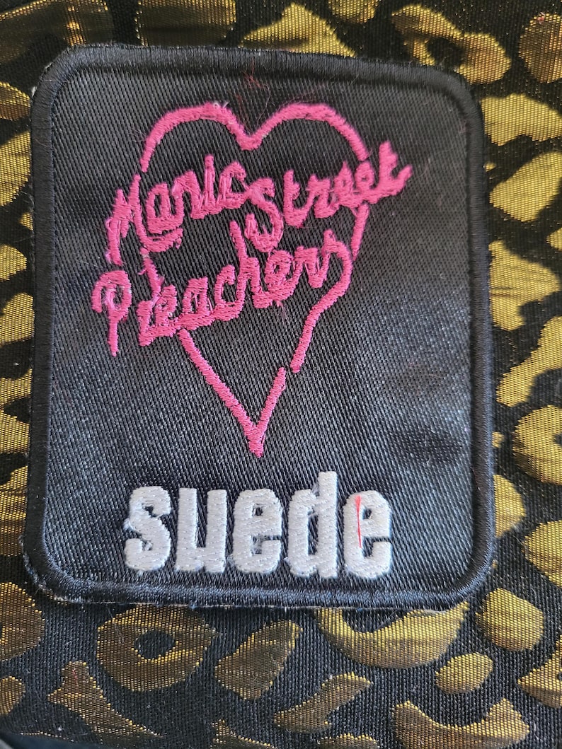 May include: A rectangular black fabric patch featuring an embroidered pink heart outline containing the text Manic Street Preachers in pink script. Below the heart, the word suede is embroidered in white block letters. The patch is on a gold leopard print.