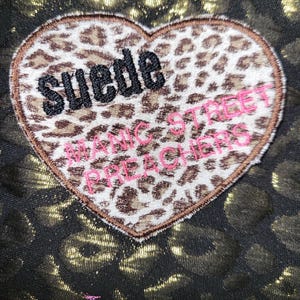 May include: A heart-shaped patch with a brown and white leopard print pattern. The word Suede is embroidered in black thread, and the text Manic Street Preachers is embroidered in pink thread. The patch is set against a black and gold leopard print fabric.