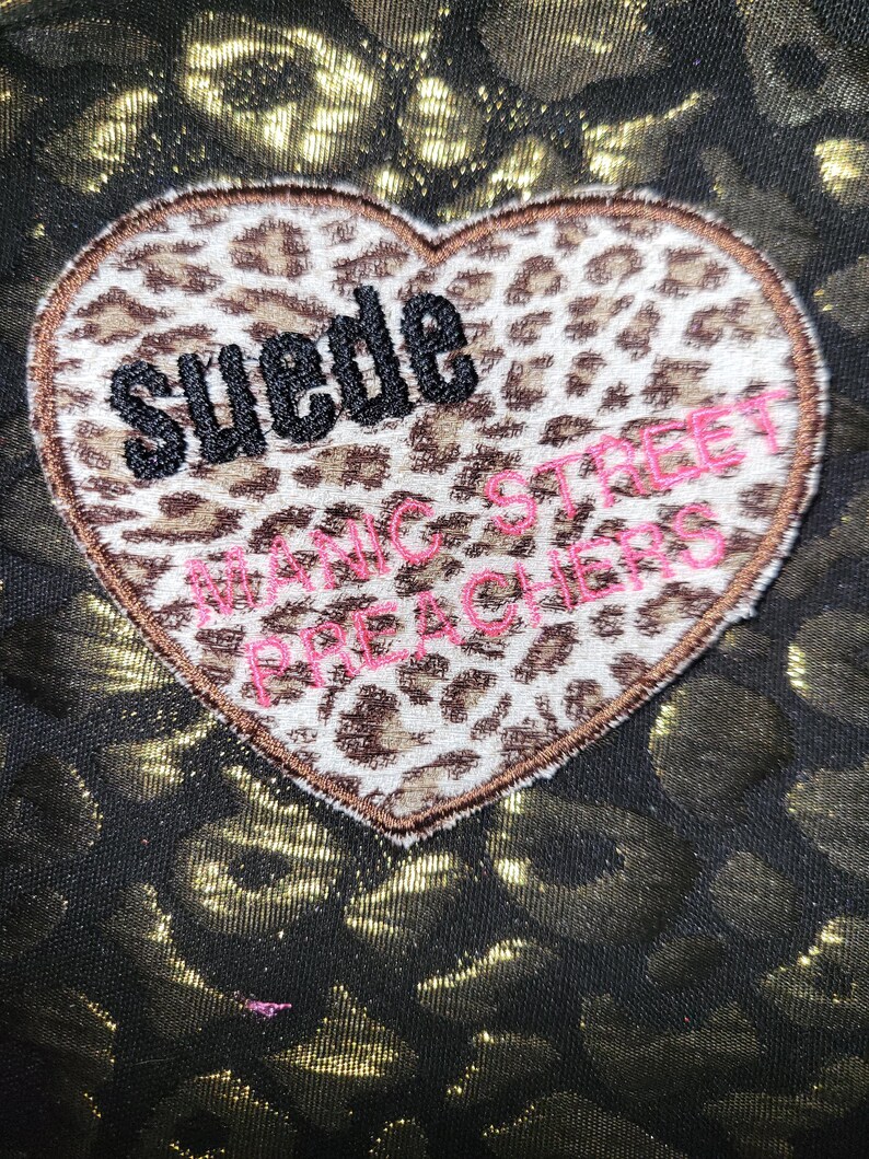 Puede incluir: Un parche en forma de coraz&oacute;n con un estampado de leopardo en marr&oacute;n y blanco. La palabra Suede est&aacute; bordada en hilo negro y el texto Manic Street Preachers est&aacute; bordado en hilo rosa. El parche est&aacute; sobre una tela con estampado de leopardo negro y dorado.