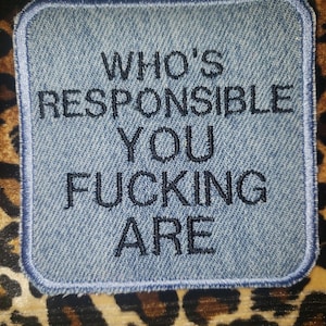 Puede incluir: Un parche de mezclilla con un borde azul y texto bordado en negro que dice "WHO'S RESPONSIBLE YOU FUCKING ARE". El parche está sobre un fondo con estampado de leopardo.