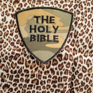 May include: Close-up of a leopard print fabric with a shield-shaped patch. The patch features the text "THE HOLY BIBLE" in black embroidery against a camouflage background. The fabric has a pattern of brown spots on a cream background.