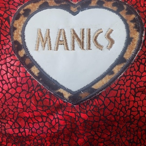 May include: Red fabric with a textured pattern, featuring a heart-shaped patch. The patch has a white centre with the word "MANICS" embroidered in gold thread. The heart is outlined with a leopard print border.