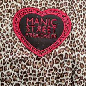 Könnte beinhalten: Nahaufnahme eines Stoffes mit Leopardenmuster. Ein herzförmiger Aufnäher mit den Worten "MANIC STREET PREACHERS" in Rot und Schwarz ist auf den Stoff genäht. Der Aufnäher hat einen roten Rand.