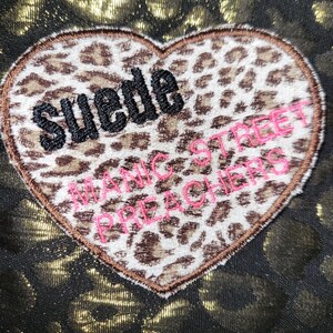 Puede incluir: Un parche en forma de coraz&oacute;n con estampado de leopardo que presenta el texto bordado en negro "Suede" y el texto bordado en rosa "Manic Street Preachers" sobre un fondo de tela oscura con un dise&ntilde;o de estampado de leopardo dorado met&aacute;lico.