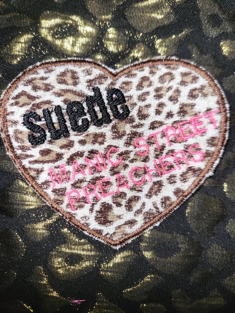 Puede incluir: Un parche en forma de coraz&oacute;n con estampado de leopardo que presenta el texto bordado en negro "Suede" y el texto bordado en rosa "Manic Street Preachers" sobre un fondo de tela oscura con un dise&ntilde;o de estampado de leopardo dorado met&aacute;lico.