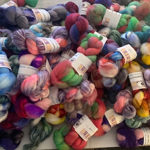 May include: A large pile of colorful yarn skeins in various shades of blue, pink, green, purple, and yellow. Each skein is wrapped with a label. The yarn is arranged on a white surface.