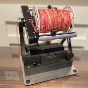 May include: A red, white, and orange yarn is wound on a white spool. The spool is attached to a black and silver yarn winder.