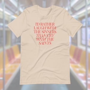 Peut inclure: Un t-shirt beige avec le texte "I'd rather laugh with the sinners than cry with the saints" imprimé en rouge.