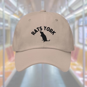 Peut inclure: Une casquette de baseball beige avec une broderie noire qui dit "Rats York" et représente la silhouette d'un rat.
