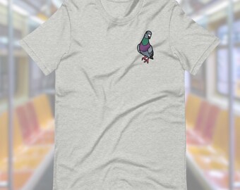 Puede incluir: Una camiseta gris jaspeada con un dibujo de una paloma de dibujos animados bordado en el pecho izquierdo. La paloma es gris con el pecho morado y las patas rojas.