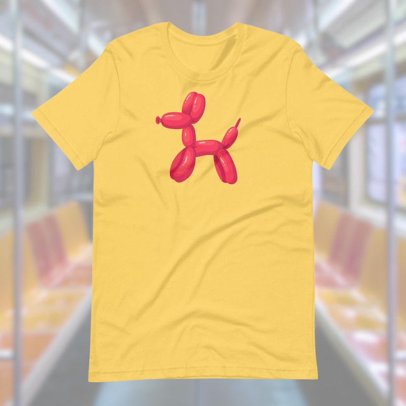 Puede incluir: Una camiseta amarilla de manga corta con un animal de globo rojo con forma de perro en la parte delantera.