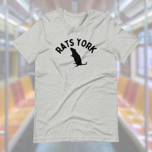 May include: A light gray t-shirt with the words "RATS YORK" in a curved font above a black silhouette of a rat. The shirt is short-sleeved and has a crew neck.