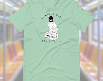 May include: A light green t-shirt with a black and white illustration of a cat wearing a mask and sitting on top of a stack of LaCroix boxes. The text "Bushwick, Baby" is above the cat and "Brooklyn, NY" is below the cat.